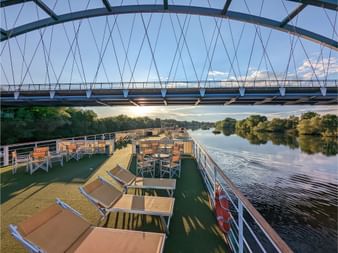 Sonnendeck der MS Casanova mit Liegestühlen und Tischen mit Blick auf einen ruhigen Fluss. Moderne Brücke überspannt den Fluss, Bäume säumen beide Ufer.