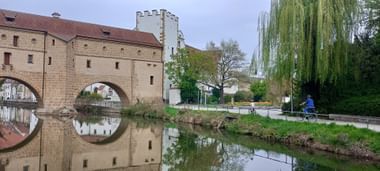 Steinernes Brückentor mit Bögen spiegelt sich im ruhigen Wasser in Amberg. Ein Radfahrer fährt am Uferweg unter einer Trauerweide.