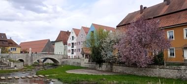 Steinbrücke über einen Fluss in Berching mit bunten historischen Gebäuden und blühenden Bäumen am Flussufer unter bewölktem Himmel.