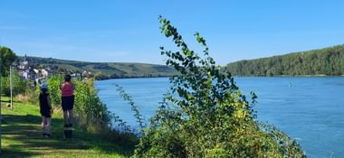 Two cyclists standing on grassy riverbank overlooking the Rhine River with green hills and village in background under blue sky.