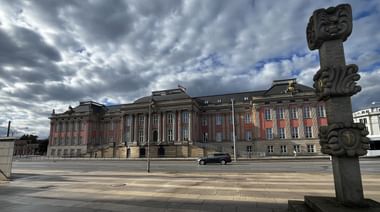 Berliner Stadtschloss mit roter und beiger Fassade unter bewölktem Himmel. Eine dekorative Steinsäule steht im Vordergrund auf einem leeren Platz.