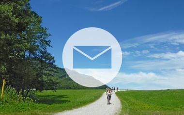 Large white envelope icon overlaid on scenic countryside with cyclists on gravel path through green fields under blue sky with trees.
