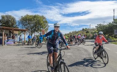 Große geführte Radgruppe auf asphaltierter Straße unter blauem Himmel. Führender Radfahrer in blauer Jacke winkt beim Fahren, viele Teilnehmer folgen.