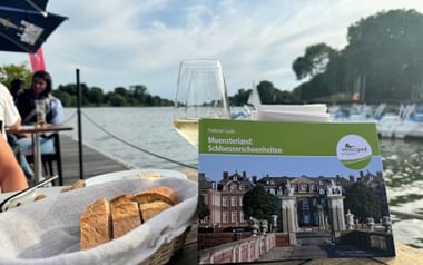Münsterland Schlösserführer auf Holztisch mit Weinglas und Brotkorb, mit Blick auf einen See mit Booten und Bäumen im Hintergrund.