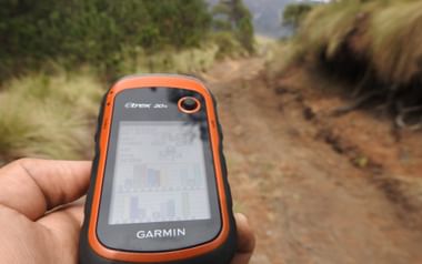 Hand hält orange-schwarzes Garmin eTrex 20x GPS-Gerät mit Kartenanzeige auf einem Feldweg mit grüner Vegetation und Hügeln im Hintergrund.