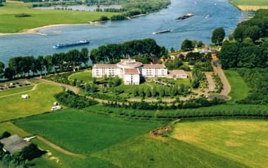 Luftaufnahme des Welcome Hotel Wesel, ein weißes mehrstöckiges Gebäude umgeben von grünen Rasenflächen und Bäumen nahe dem Rhein mit Frachtschiffen.