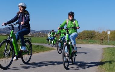 Gruppe von Radfahrern auf grünen E-Bikes auf einem asphaltierten Radweg bei Marburg. Blauer Himmel, grüne Felder und Hügel im Hintergrund.