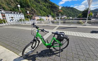 Grünes E-Bike mit Gepäckträger auf gepflasterter Brücke in Bad Ems. Hügel mit Gebäuden und blauer Himmel mit Wolken im Hintergrund.