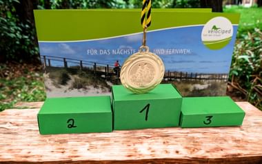 Grünes Siegertreppchen mit Plätzen 1, 2 und 3, Goldmedaille hängt über dem ersten Platz. Velociped-Banner mit Radfahrerbild im Hintergrund.