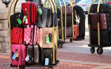 Mehrere goldene Gepäckwagen beladen mit bunten Koffern in Rot, Pink, Schwarz und Punktmustern mit Gepäckanhängern auf Ziegelpflaster.