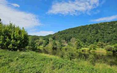 Ruhiger Fluss schlängelt sich durch üppige grüne Landschaft mit dicht bewaldeten Hügeln unter blauem Himmel mit vereinzelten Wolken.