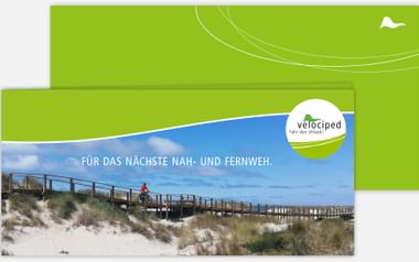 Grüner Velociped Geschenkgutschein zeigt Radfahrer auf Holzsteg durch Küstendünen mit Text 'FÜR DAS NÄCHSTE NAH- UND FERNWEH'