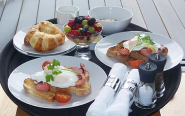 Frühstückstablett mit Croissant, frischen Beeren, Müslischale, zwei Tellern mit Spiegeleiern, Speck, Toast und Würstchen sowie Besteck.