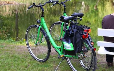 Velociped 7-speed touring bike by lake Green Velociped 7-speed touring bicycle with black panniers parked on grass near a lake, with a white bench visible in the background.
