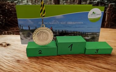 Grünes Siegerpodest mit Plätzen 1, 2 und 3. Eine Goldmedaille hängt über Platz 2. Dahinter eine Velociped-Urkunde mit Stegmotiv.