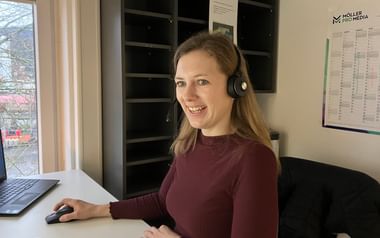 Christine sitzt an einem weißen Büroschreibtisch, trägt einen bordeauxroten Pullover und ein Headset und lächelt beim Arbeiten am Laptop.