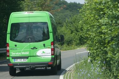 Grüner Mercedes-Benz Sprinter mit 'Service/Mobil' und Velociped-Aufschrift fährt auf einer Landstraße umgeben von grünen Bäumen.