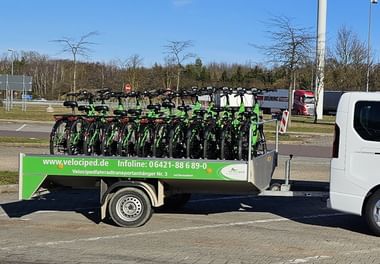 Grüner Velociped-Anhänger beladen mit mehreren Fahrrädern, an einen weißen Transporter angehängt, auf einer Straße mit Bäumen und Gebäuden im Hintergrund.