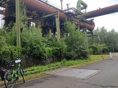 Grünes Fahrrad auf Weg neben bewachsenen Industrieanlagen mit großen Rohren im Landschaftspark Duisburg im Ruhrgebiet.
