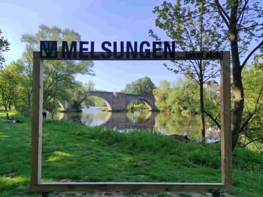 Holzrahmen mit 'Melsungen'-Schild rahmt Blick auf historische Steinbogenbrücke über die Fulda, umgeben von grünen Bäumen und Gras.