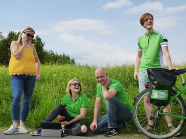 Velociped Team Ansprechpartner im Freien Vier Velociped Teammitglieder auf grüner Wiese. Frau im gelben Top telefoniert, zwei Personen in grünen Shirts sitzen, Mann mit Fahrrad.