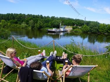 Cyclists relaxing by Weser River ferry Four cyclists relaxing in deck chairs by the Weser River in Schweringen, watching a small ferry cross the water surrounded by green countryside.