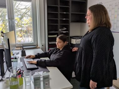 Two women in an office: Annabel seated at desk with computer and Miriam standing beside her. Office supplies and storage shelves visible.