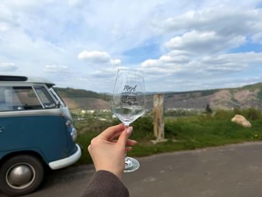 Hand hält Weinglas mit Mosel Bulli Tour Logo, Oldtimer-VW-Bus und Moseltal-Weinberge im Hintergrund unter bewölktem Himmel.