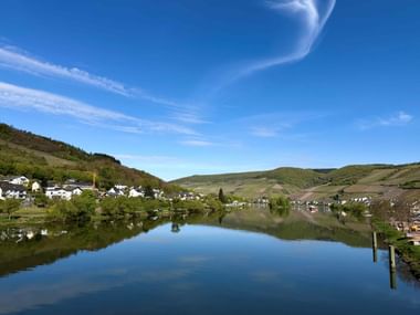 Calm Moselle River reflecting blue sky, with villages on both banks and green vineyard-covered hills under clear skies near Zell and Treis-Karden.