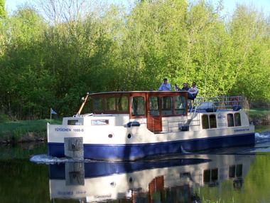 Weiß-blaues Hausboot namens Tütüchen fährt auf ruhigem Wasser mit Passagieren an Deck, umgeben von grünen Bäumen an der Havel.