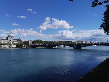 Wide view of a stone bridge crossing the Rhône River in Lyon under blue sky with white clouds. Historic buildings line the left riverbank.