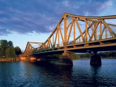 Glienicker Brücke, eine Stahlfachwerk-Brücke mit gelb-orangem Rahmen über blauem Wasser unter bewölktem Himmel zur goldenen Stunde.