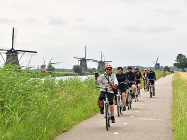 Gruppe von Radfahrern auf gepflastertem Weg durch grüne Landschaft in Kinderdijk mit traditionellen holländischen Windmühlen im Hintergrund.