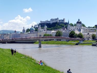 Blick auf Salzburg von der Salzach mit der historischen Altstadt, barocken Kirchen, der Festung Hohensalzburg auf dem Berg und einer Brücke.