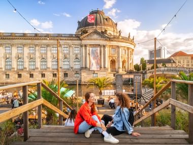 Zwei Frauen auf Holzbrücke beim Bode-Museum Zwei junge Frauen sitzen auf Holzbrückenstufen mit der historischen Barockkuppel des Bode-Museums in Berlin im Hintergrund zur goldenen Stunde.