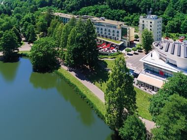 Luftaufnahme des Victors Residenz Saarbrücken Hotelkomplexes mit modernen Gebäuden, Parkplatz und Lage am See umgeben von Wald.