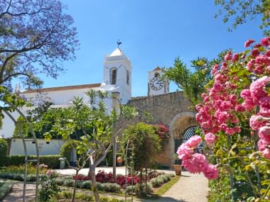 White church Igreja do Castelo in Tavira with bell tower and clock tower, surrounded by blooming pink bougainvillea and lush gardens.