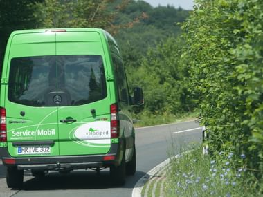 Green Mercedes-Benz Sprinter van with 'Service/Mobil' and Velociped branding driving on a country road surrounded by green trees.