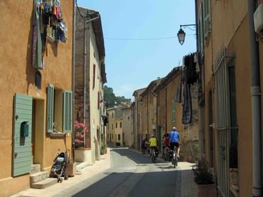 Three cyclists riding through a narrow stone street in a Provence village with ochre-colored buildings, green shutters, and laundry hanging from balconies.