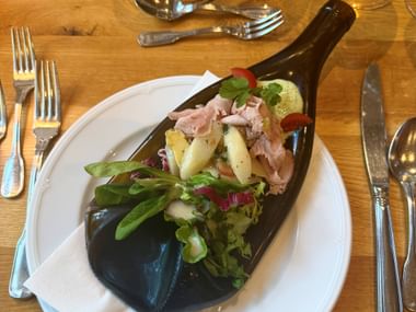 Erster Gang serviert auf einer längs geschnittenen dunklen Weinflasche mit weißem Spargel, Fleischscheiben, Kirschtomaten und frischem Grün.