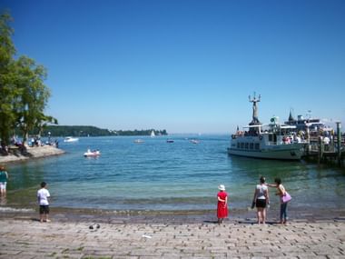 Konstanzer Hafen am Bodensee mit weißer Passagierfähre am Pier. Menschen stehen auf dem Kopfsteinpflaster-Ufer und beobachten Boote.