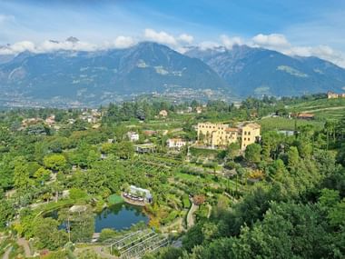 Aerial view of Trauttmansdorff Castle surrounded by botanical gardens with terraced landscapes, pond, and Alpine mountains in background.