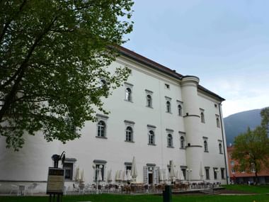 Weißes Renaissance-Schloss Porcia in Spittal mit zylindrischen Türmen, Bogenfenstern und Café-Bestuhlung auf dem Rasen unter grünen Bäumen.
