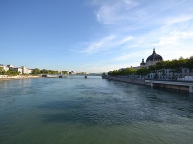 Wide view of the Rhône River in Lyon with tree-lined banks, a domed church on the right, bridges in the distance, and clear blue sky above.
