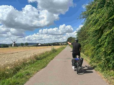 Cyclist with panniers riding on paved path between wheat field and green hedge, with windmill visible in distance under blue sky with clouds.