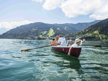 Red rowing boat with five people on turquoise Zeller See. Mountains and green slopes visible in background under partly cloudy sky.