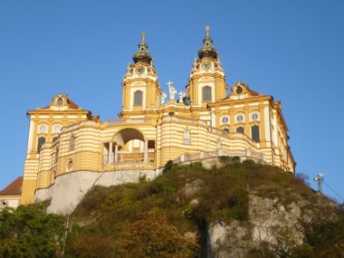 Stift Melk auf Hügel in Österreich Gelbes Barockkloster mit Zwillingstürmen und grünen Kuppeln auf einem felsigen Hügel vor klarem blauen Himmel, umgeben von Vegetation.