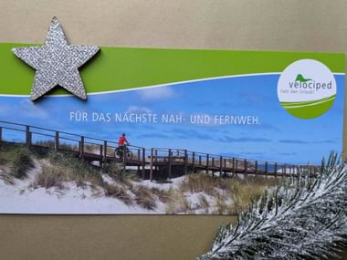 Velociped gift voucher showing cyclist on wooden boardwalk at beach, decorated with silver star and pine branch. German text reads 'For the next near and far'.