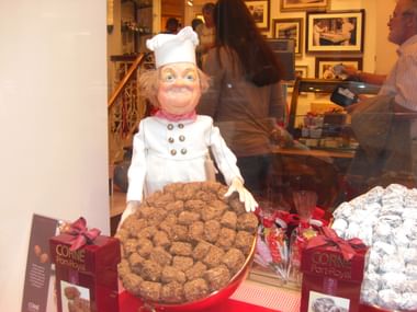 Dekorative Kochfigur in weißer Uniform und Mütze präsentiert Schokoladentrüffel in einem belgischen Schokoladenladen mit Kunden im Hintergrund.