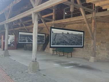 Überdachte Holzkonstruktion in der Zeche Nachtigall mit historischen Fotografien und Grubenwagen. Holzbalken tragen das Dach über gepflastertem Bereich.